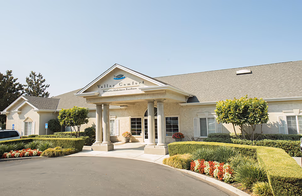 Exterior view of Valley Comfort, a single-story building with a covered entrance supported by columns. The building is surrounded by neatly trimmed bushes, trees, and flower beds under a clear blue sky.