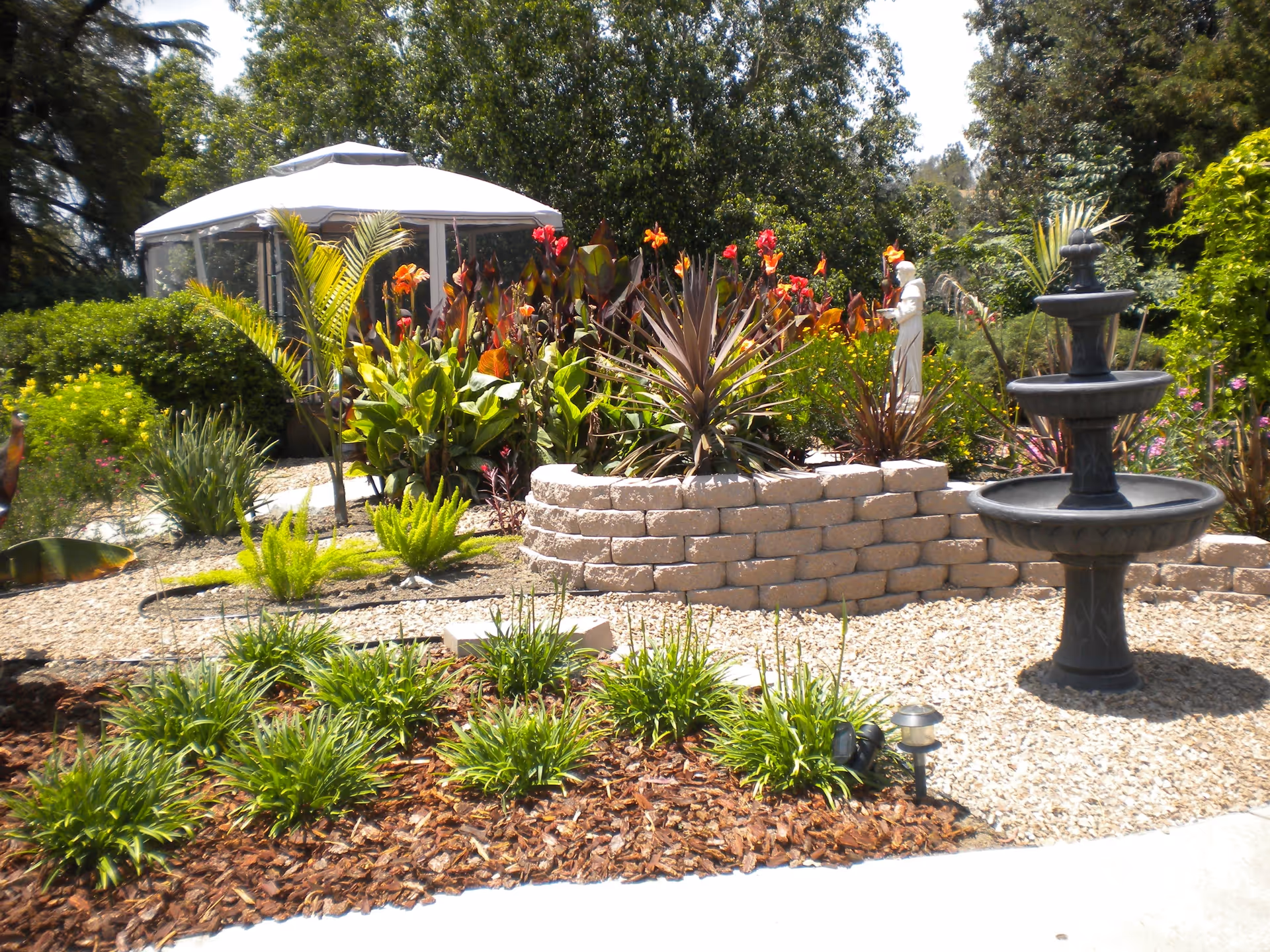 A vibrant garden area with various green plants and colorful flowers surrounding a tiered black water fountain. There is a white gazebo in the background and a statue among the plants. The garden features a stone retaining wall and gravel pathways.