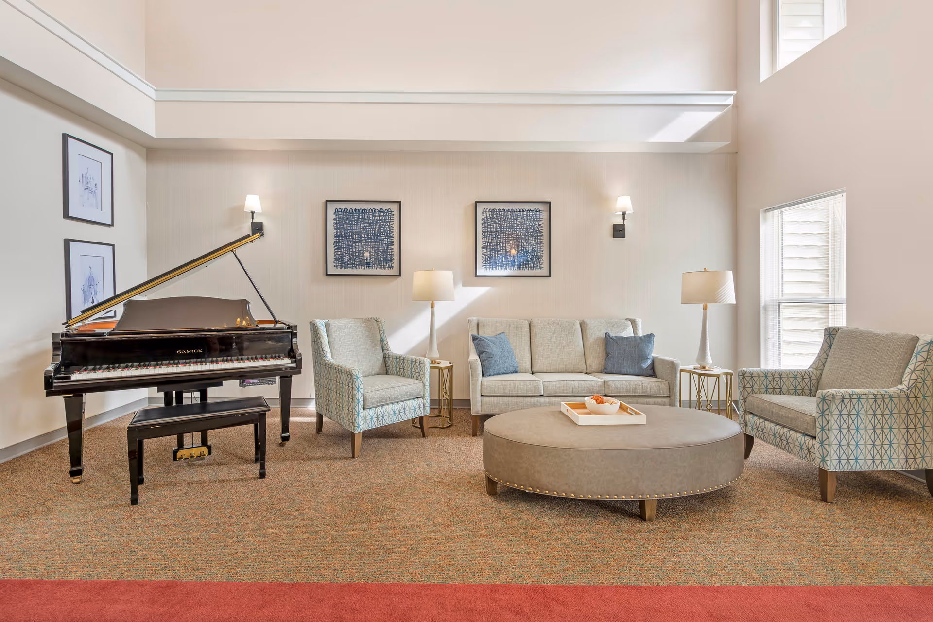 A bright and spacious living room with a grand piano on the left, two patterned armchairs, a beige sofa with blue cushions, two side tables with lamps, a large round ottoman with a tray, and two framed abstract artworks on the wall.
