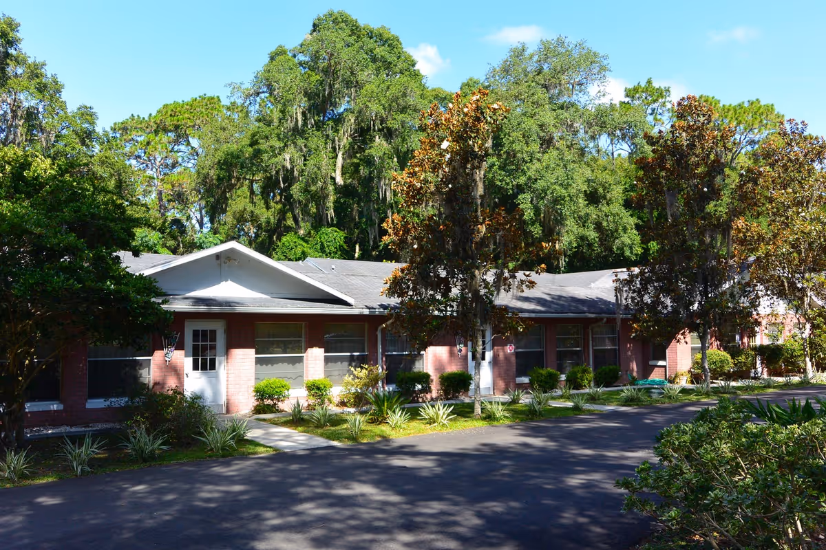 Exterior view of a single-story brick building with multiple windows and white doors, surrounded by well-maintained landscaping including bushes, small plants, and trees, under a clear blue sky.