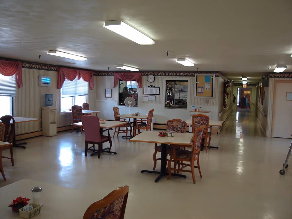 A dining room in a senior living facility with several tables and chairs arranged neatly. The room has large windows with pink curtains, fluorescent ceiling lights, and a hallway leading to other areas. There are small decorations on the tables and a clock on the far wall above a service window.