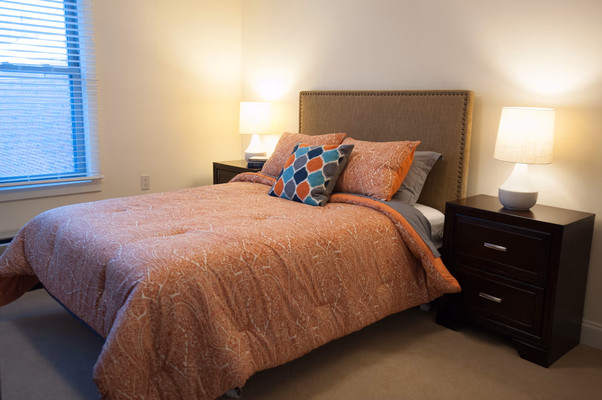 A cozy bedroom with a neatly made bed featuring an orange patterned comforter and multiple pillows, including a decorative one with a colorful geometric pattern. There are two dark wooden nightstands on either side of the bed, each with a lit table lamp. A window with blinds is visible on the left side of the room.