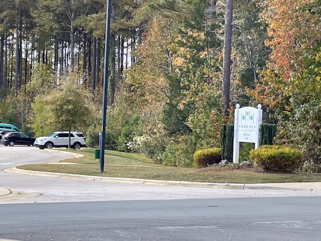 View of a parking lot area with several parked cars next to a landscaped area with trees and bushes. A white sign with the text 'Cadence Garner' is visible near the edge of the parking lot, surrounded by greenery.