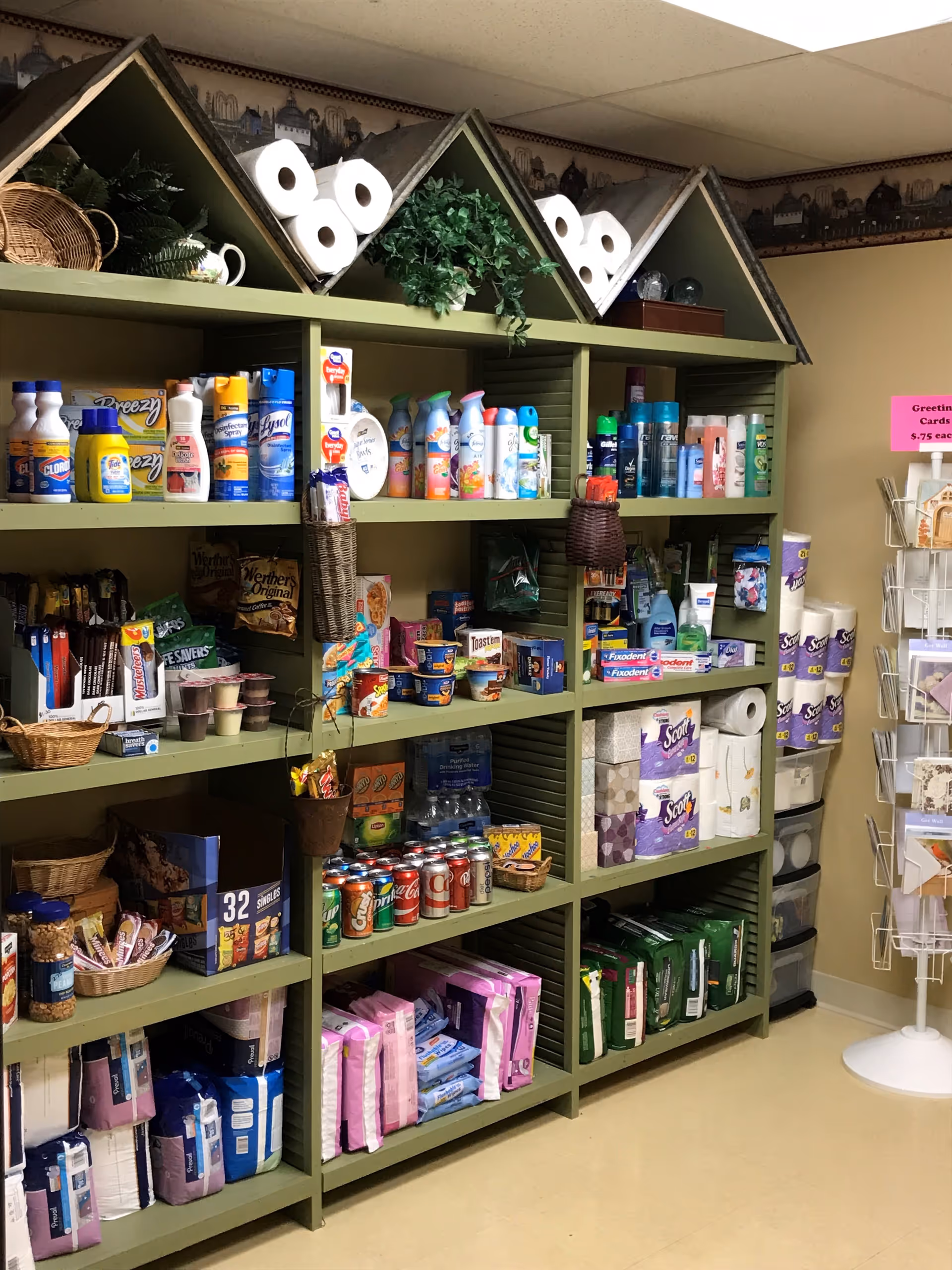 Shelves stocked with various household and personal care items including cleaning supplies, air fresheners, snacks, canned sodas, paper towels, and adult incontinence products in a well-lit room with a beige floor and walls.