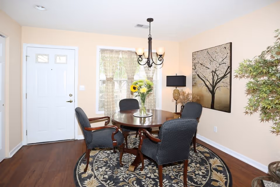 A dining area with a round wooden table surrounded by four upholstered chairs on a patterned circular rug. A vase with sunflowers and other flowers is on the table. There is a window with sheer curtains behind the table, a chandelier hanging above, a door to the left, a floor lamp and decorative wall art of a tree on the right wall, and a potted plant in the corner.