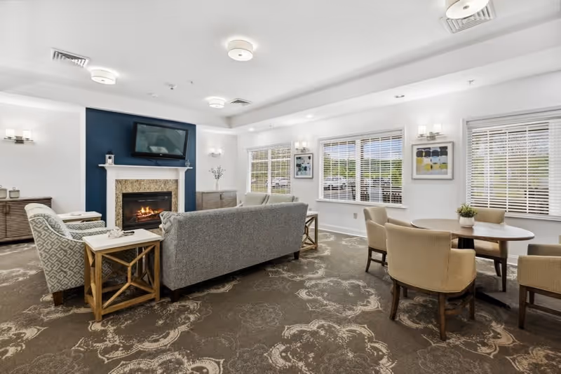 A bright and spacious living room area with a patterned carpet, a gray sofa, two armchairs, a fireplace with a TV mounted above it, and a round table with four beige chairs near windows with white blinds. The walls are white with modern light fixtures and framed artwork.