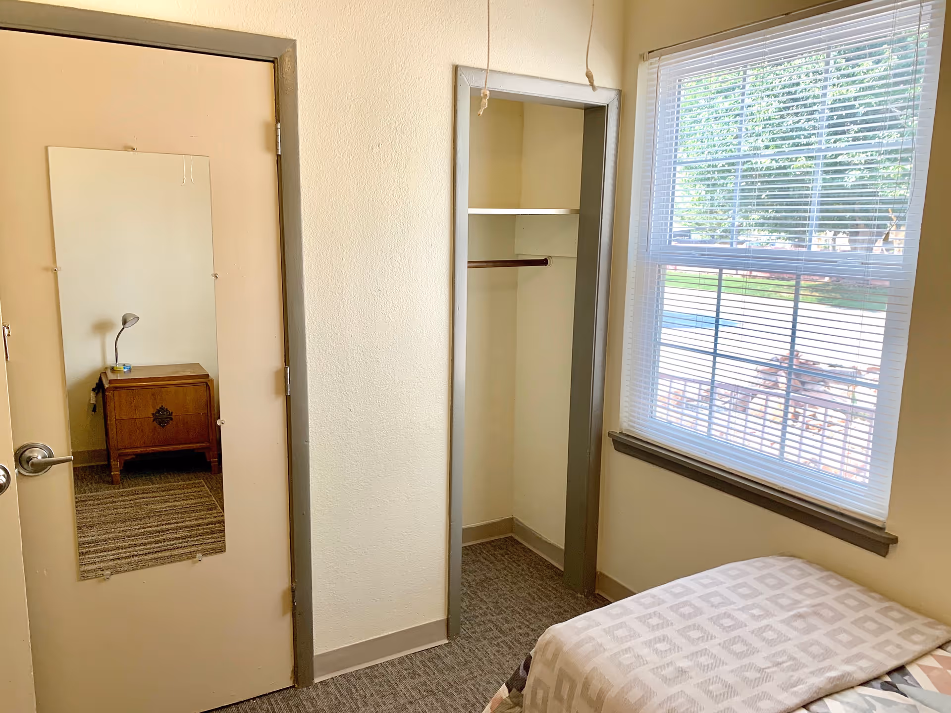 A small bedroom with a bed partially visible on the right side covered with a patterned blanket. There is a window with blinds letting in natural light. An open closet with a hanging rod and shelf is in the center. A door on the left has a full-length mirror attached, reflecting a wooden nightstand with a small lamp and an item on top. The walls are light-colored and the floor is carpeted.