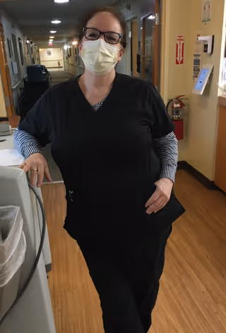 A woman wearing black scrubs, a striped long-sleeve shirt underneath, glasses, and a face mask stands in a hallway of a senior living facility, leaning against a piece of equipment. The hallway has wooden flooring and is lined with doors and handrails.