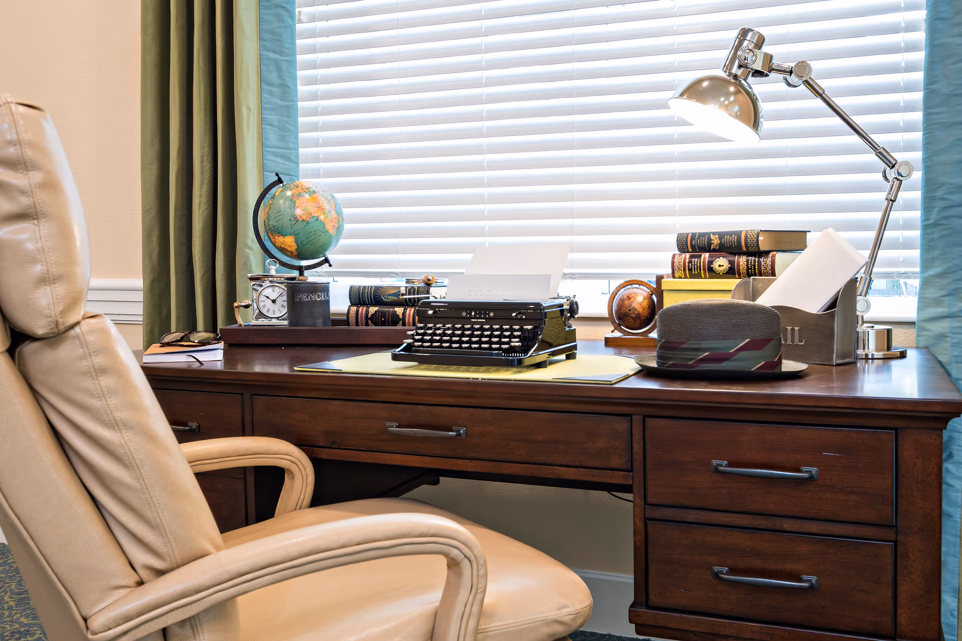 A wooden desk with a beige leather office chair in front. On the desk are a vintage typewriter with a sheet of paper, a globe, a small clock, several stacked books, a desk lamp, a hat, and a container holding envelopes. The background features a window with white blinds and green curtains.