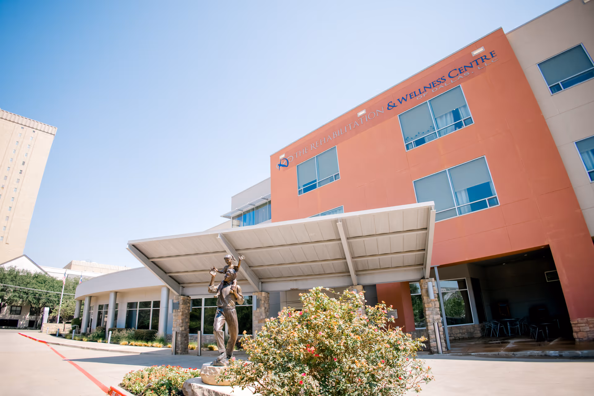 Exterior view of The Rehabilitation & Wellness Centre of Dallas building with a covered entrance and a bronze statue of an adult carrying a child on their shoulders in front of the entrance. There are bushes with flowers near the statue and clear blue sky above.