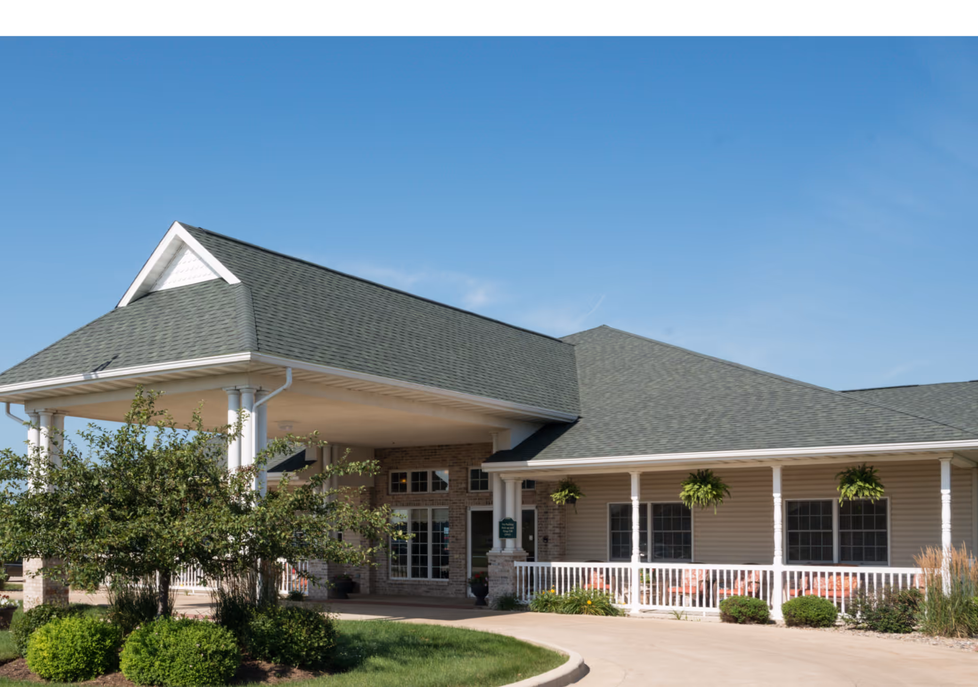 Exterior view of The Glenwood Supportive Living of Mt. Zion building with a covered entrance, green roof, white pillars, and a well-maintained garden with bushes and hanging plants under a clear blue sky.