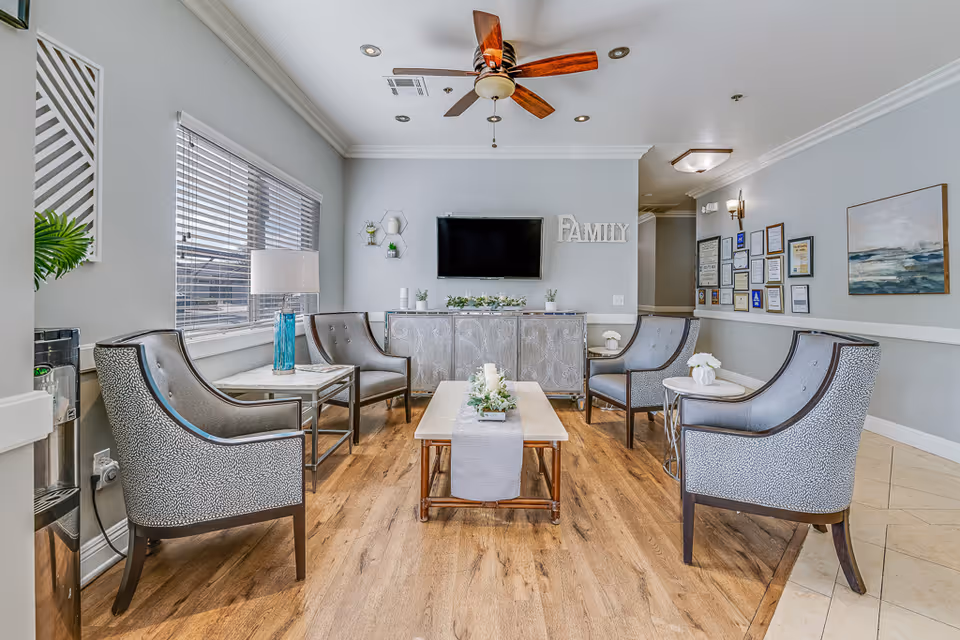 A cozy living room area with four patterned armchairs arranged around a wooden coffee table with a decorative runner and candles. A side table with a blue glass lamp is positioned near a window with blinds. A ceiling fan with wooden blades is mounted on the white ceiling. On the far wall, a flat-screen TV is mounted above a decorative cabinet, with the word 'FAMILY' displayed beside it. The walls are painted light gray and adorned with framed certificates and artwork.