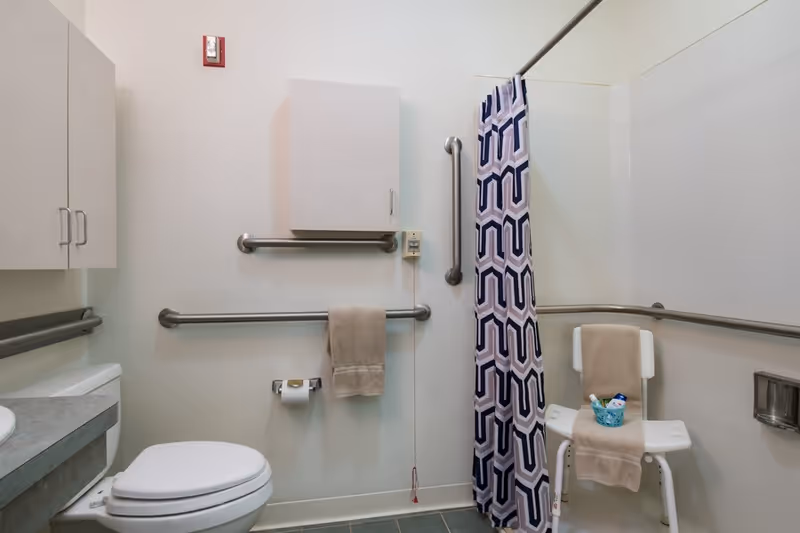 A clean, accessible bathroom with a toilet, grab bars on the walls, a shower area with a patterned curtain, and a white shower chair holding a towel and toiletries. There are cabinets mounted on the wall and a towel hanging on a grab bar.