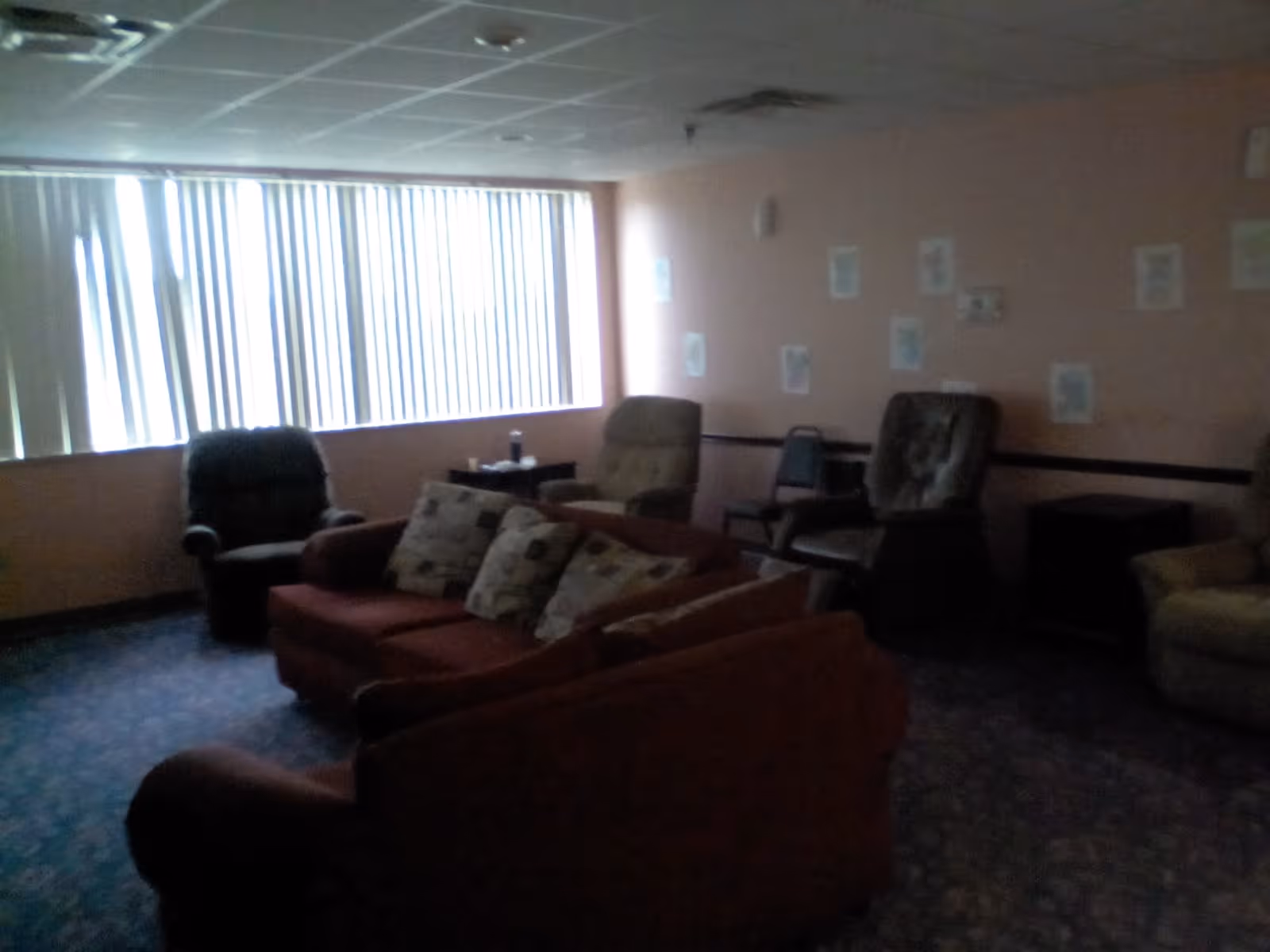 A dimly lit living room with several cushioned armchairs and a sofa with patterned pillows. The room has a large window with vertical blinds partially closed, and light-colored walls with some papers or decorations taped to them.