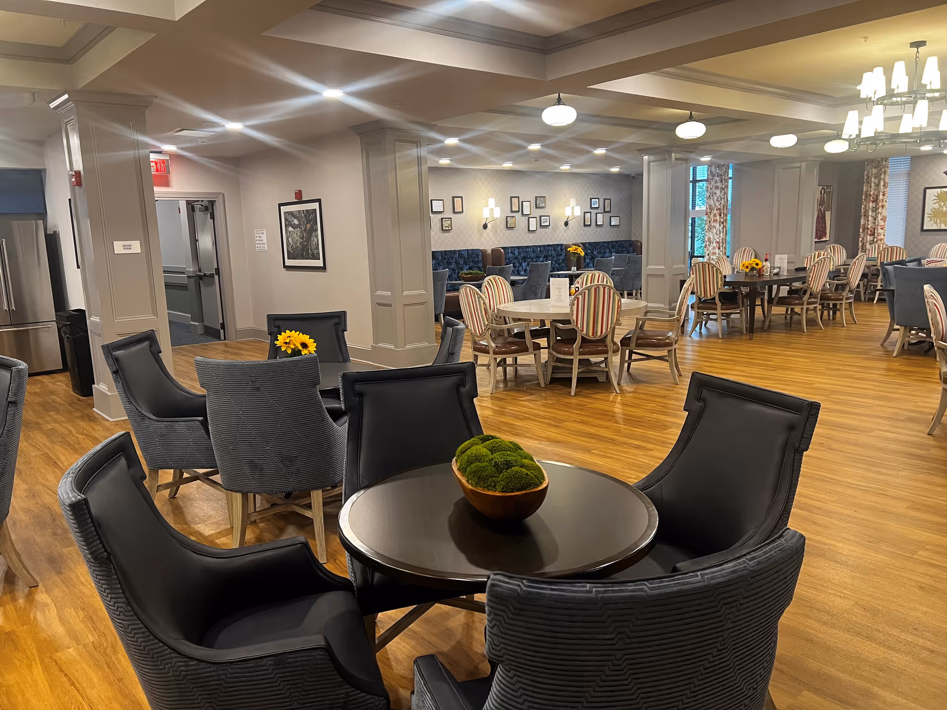 A spacious dining room in a senior living facility with multiple round and rectangular tables surrounded by various upholstered chairs. The room features wood flooring, soft lighting from ceiling fixtures and chandeliers, decorative wall art, and floral arrangements on some tables. There are columns and large windows with patterned curtains in the background.