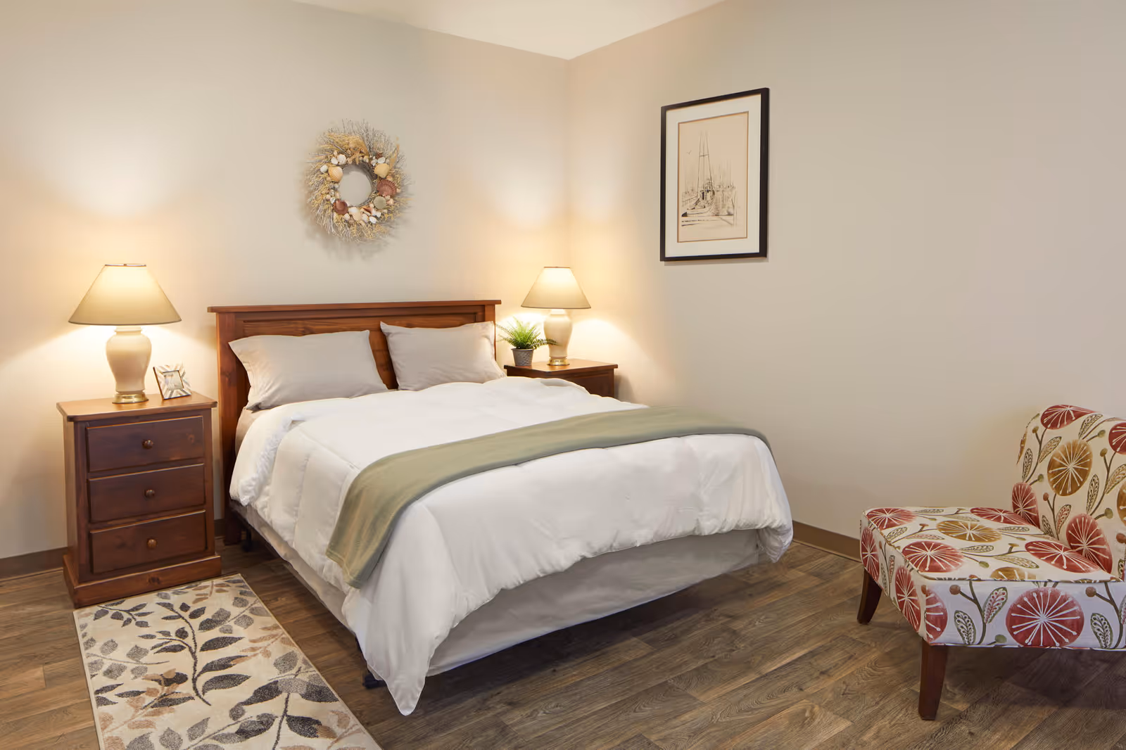 Well-lit bedroom with a made double bed flanked by wooden nightstands and lamps, a patterned armchair, and simple wall decor.