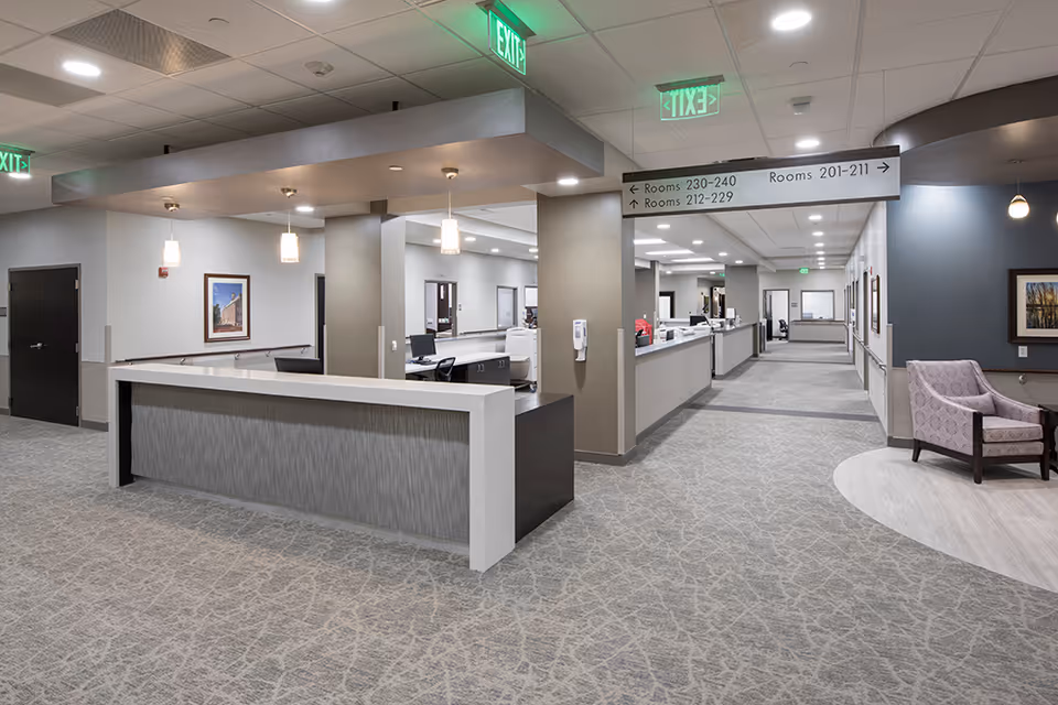 Spacious modern reception desk and long hallway in a senior living facility with seating, overhead room signage, and soft lighting.