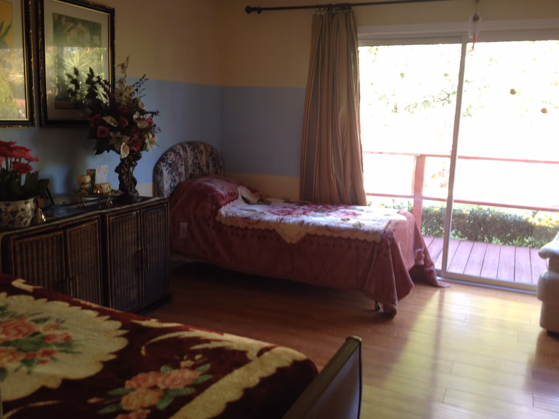 A bedroom with two single beds covered in floral blankets. There is a wooden cabinet with a vase of flowers and framed pictures on the wall above it. A large sliding glass door with beige curtains opens to an outdoor balcony with greenery visible outside.
