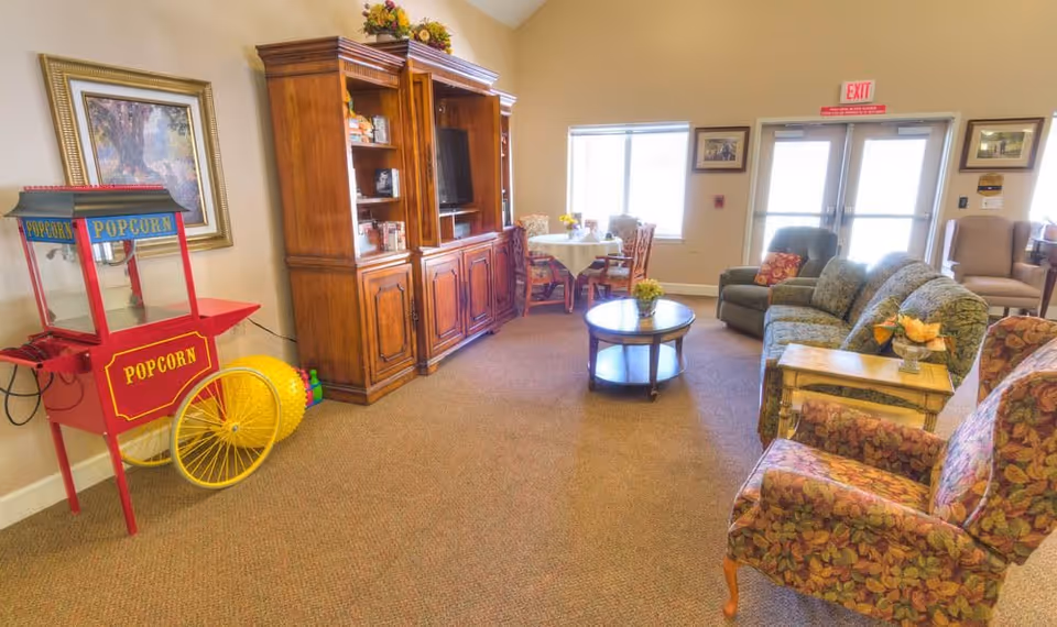 A cozy living room area in a senior living facility with floral upholstered armchairs, a green patterned sofa, a wooden coffee table with a small flower arrangement, and a wooden entertainment center with a TV. There is a red popcorn machine on the left side and a small round table with chairs near the window. The room has beige walls and carpeted floor, with an exit door and windows letting in natural light.