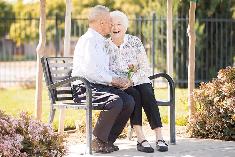 An elderly couple sitting on a metal bench outdoors in a garden area. The man is kissing the woman on the cheek while holding her hands, and the woman is smiling and holding a small bouquet of flowers. There are plants and trees around them and a metal fence in the background.
