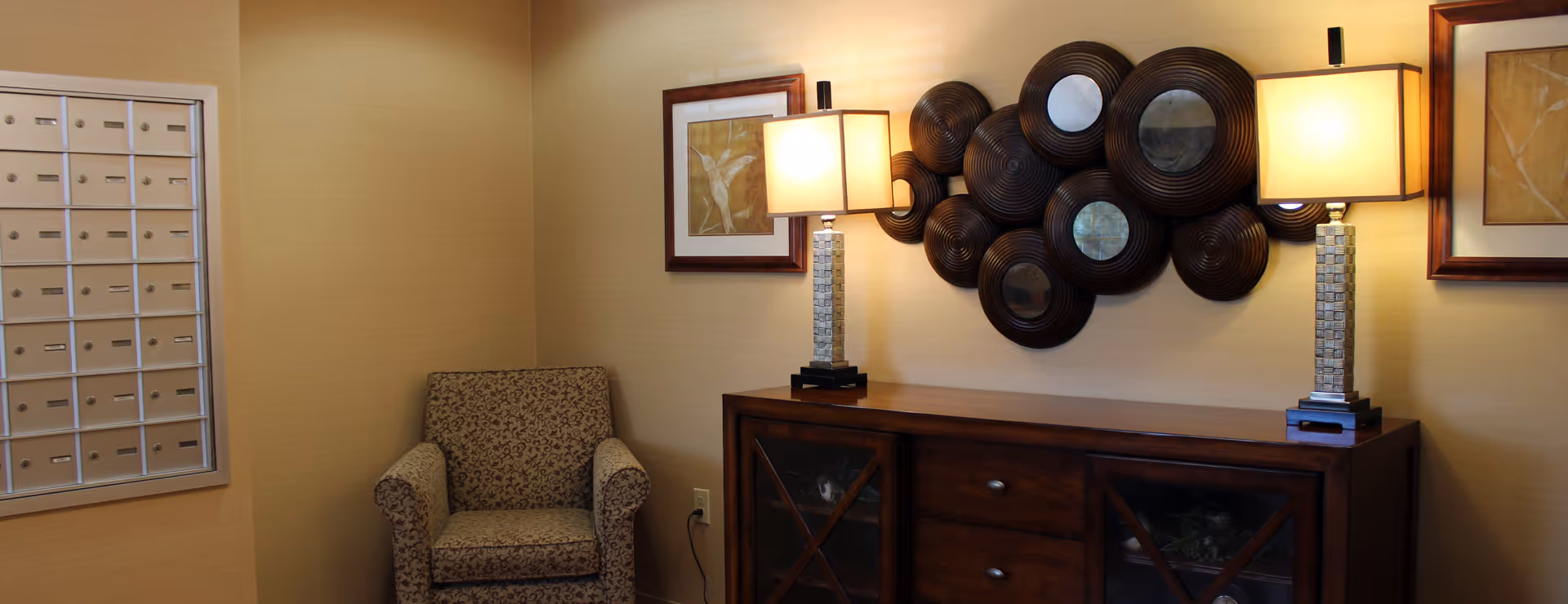 A cozy interior corner of a senior living facility featuring a patterned armchair, a wooden cabinet with glass doors, two table lamps with square shades, decorative circular wall mirrors, and framed artwork on beige walls. A set of mailboxes is mounted on the left wall.