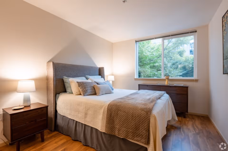 A cozy bedroom with a large bed featuring a gray upholstered headboard, multiple pillows, and a beige quilted blanket. There are two wooden nightstands with lamps on either side of the bed. A large window shows green trees outside, and a wooden dresser is placed beneath the window. The room has light-colored walls and wooden flooring.