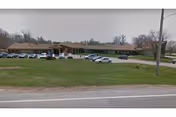 Exterior view of Leonardville Nursing Home, a single-story building with a brown roof and brick facade, surrounded by a grassy area and a parking lot with several cars parked.