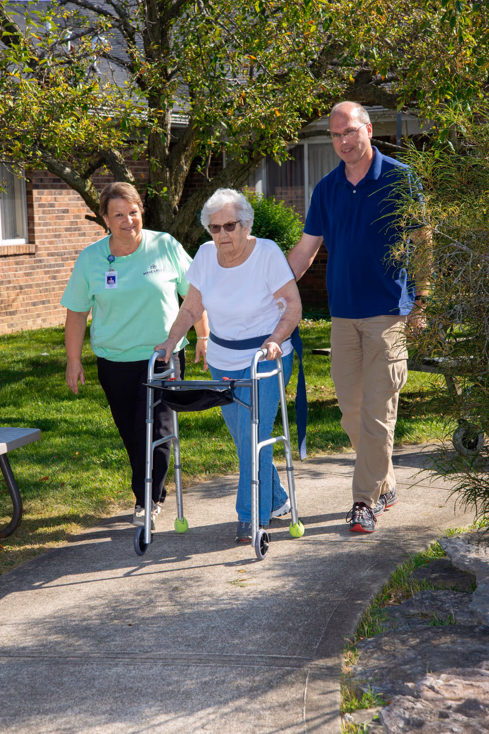 An elderly woman using a walker is being assisted by a woman in a green shirt and a man in a blue shirt as they walk on a paved path outside near a brick building with trees and greenery around.