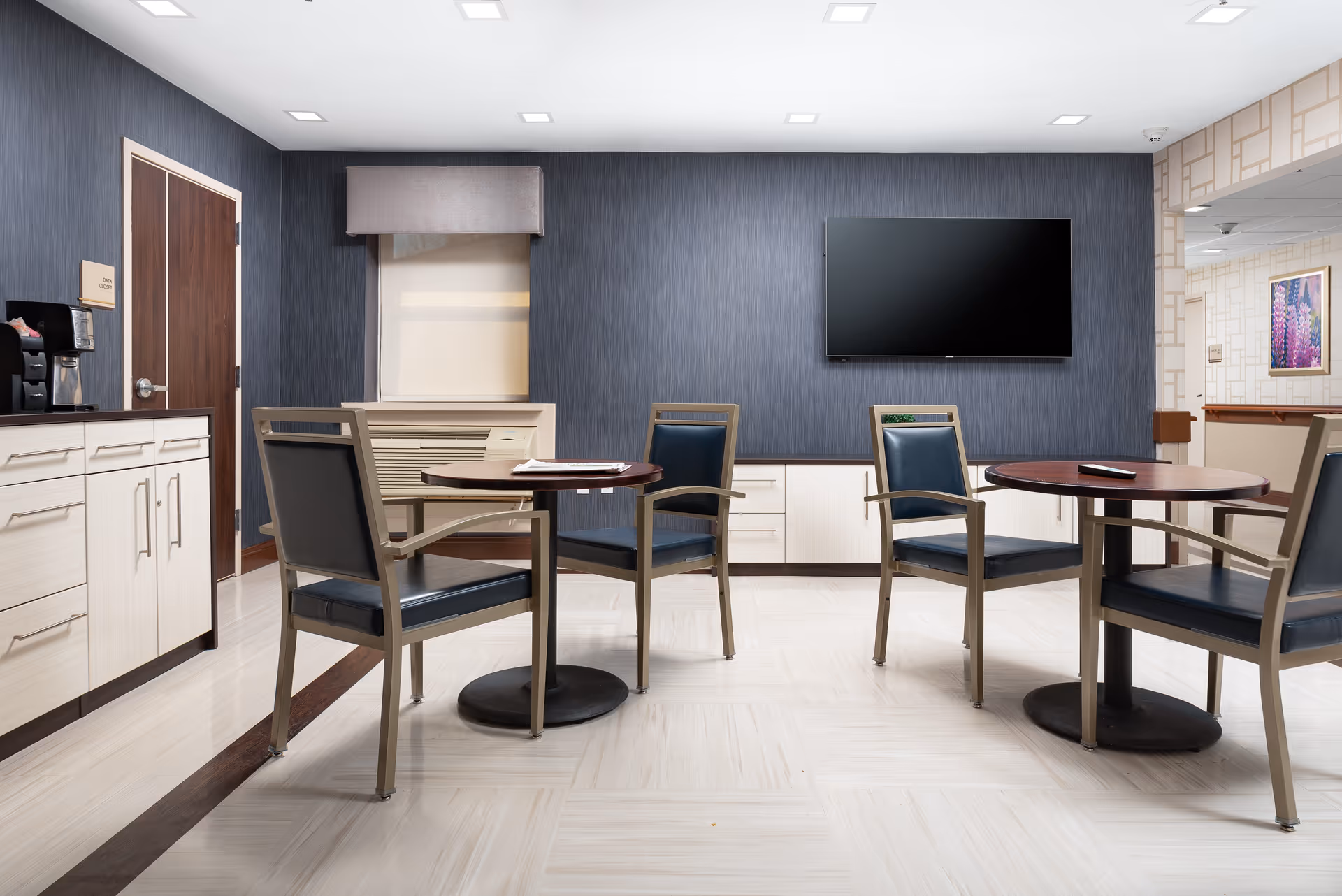 A clean and modern common area with two round tables, each surrounded by three chairs with dark blue cushions and metal frames. The room has light-colored tiled flooring, a dark blue textured accent wall with a large flat-screen TV mounted on it, and light cabinetry along the walls. There is a coffee machine on the left countertop and a window with a beige valance above an air conditioning unit. The ceiling has recessed lighting, and an open doorway on the right leads to another area with patterned wallpaper and a framed floral picture.