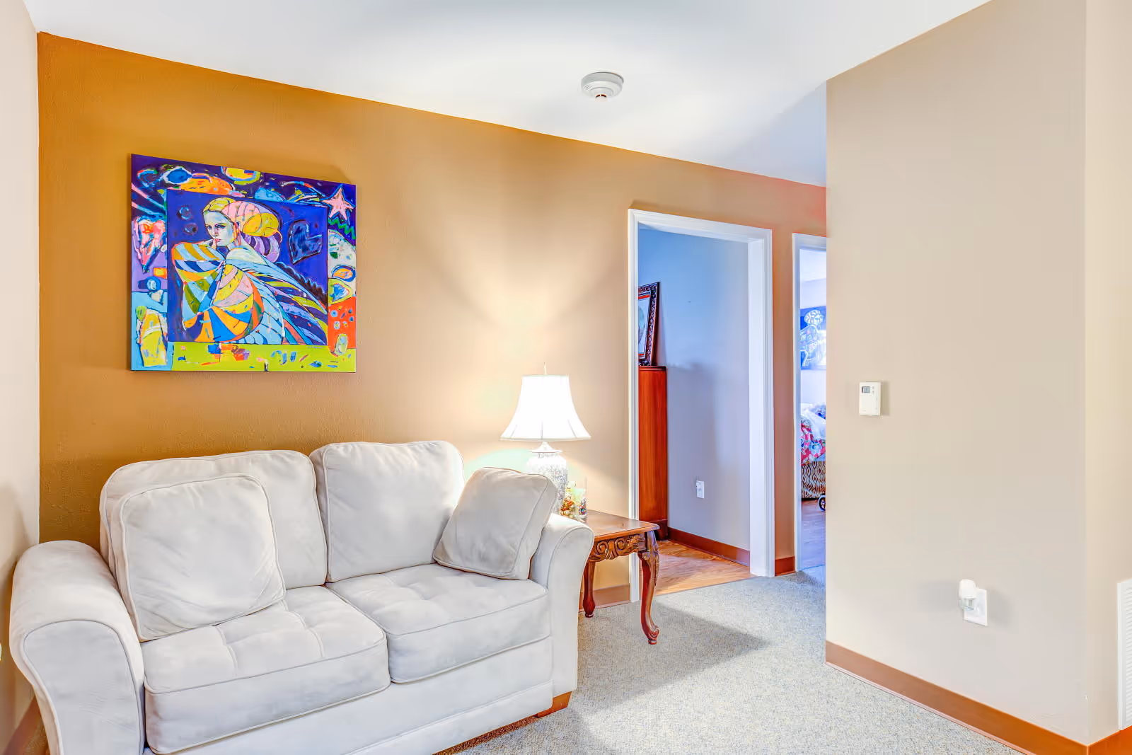 A cozy living room area with a white cushioned sofa against a warm orange accent wall. Above the sofa hangs a colorful abstract painting. Next to the sofa is a wooden side table with a decorative lamp. Two doorways lead to other rooms, one showing a glimpse of a bed with a colorful bedspread.
