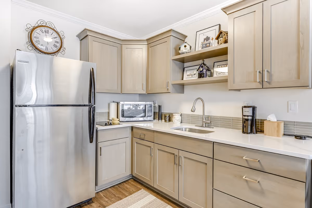 Modern kitchen with light wood cabinets, a stainless steel refrigerator, a microwave, a sink with a faucet, a coffee maker, and decorative items including framed pictures and birdhouses on open shelves.