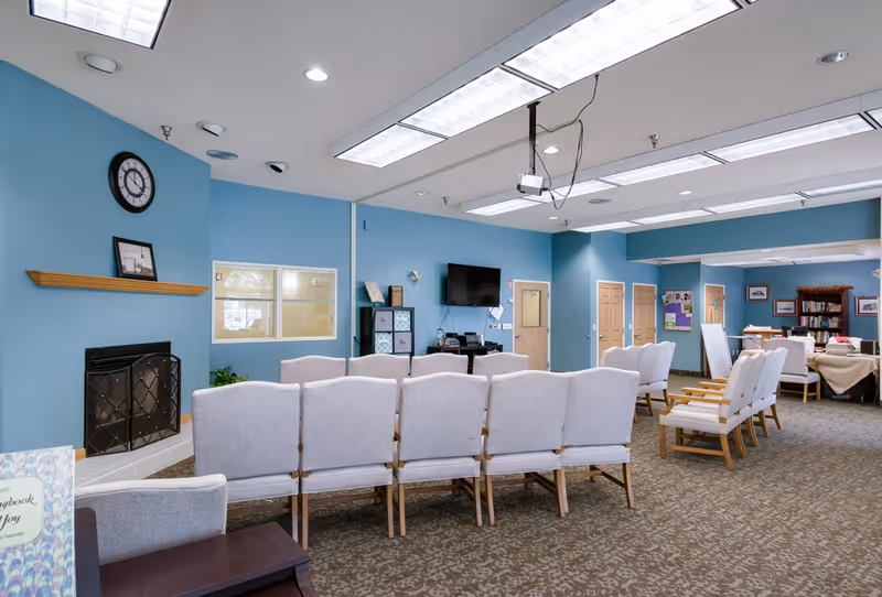 Interior view of a senior living facility common area with rows of light gray cushioned chairs facing a wall-mounted TV. The walls are painted blue, and there is a fireplace with a clock and framed picture above it. The room has fluorescent ceiling lights and carpeted floors. In the background, there are tables with chairs and bookshelves.