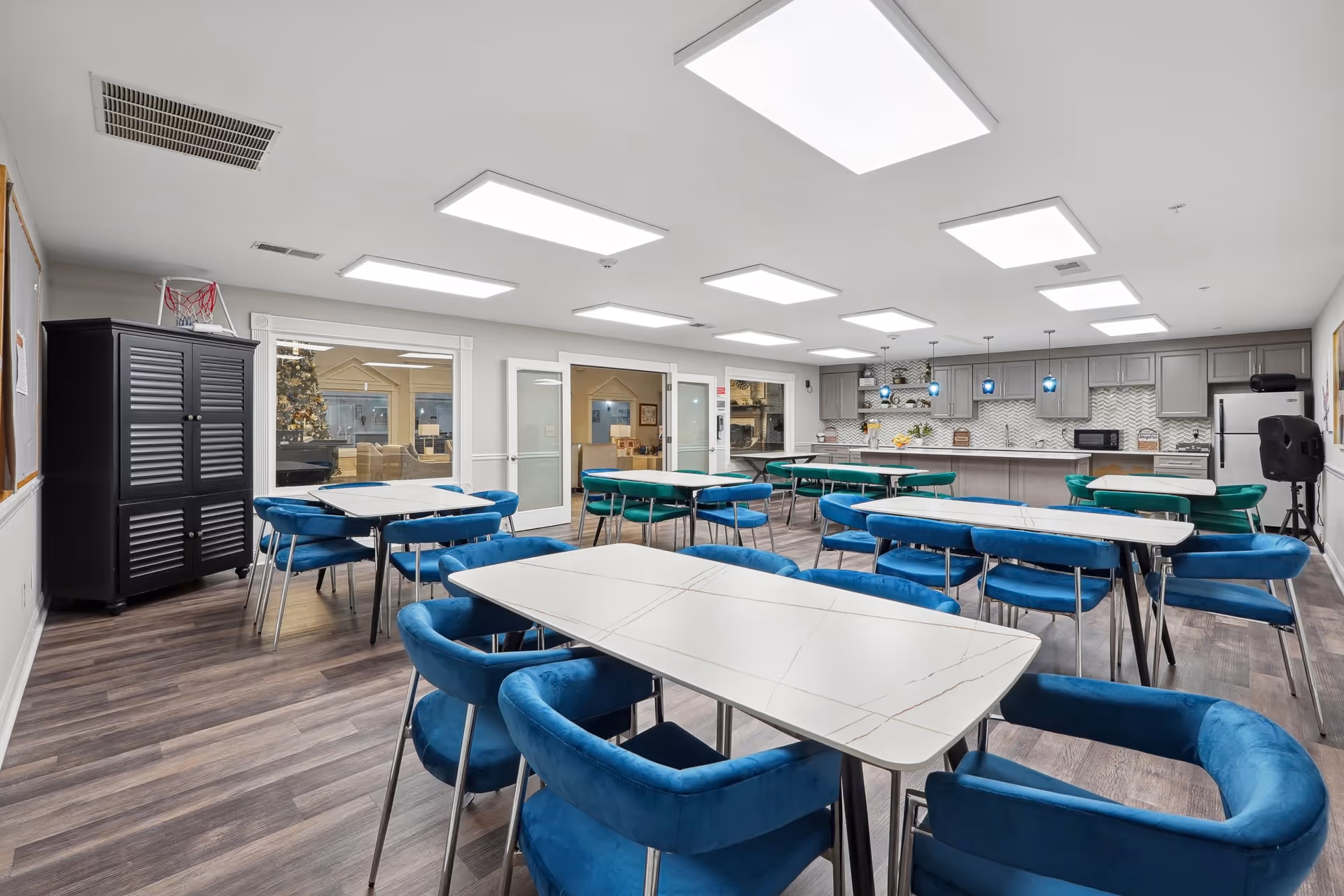 A spacious, well-lit dining area with multiple white tables and blue cushioned chairs arranged neatly. The room features modern ceiling lights, wood flooring, and a kitchen area with gray cabinets, a refrigerator, microwave, and a long counter with pendant lights. There is a black cabinet and a bulletin board on the left wall, and large windows and glass doors leading to another room.