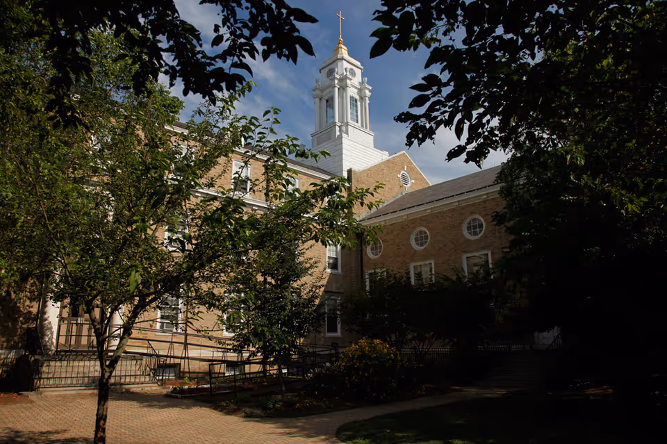 Exterior view of a brick building with a white steeple topped with a gold cross, partially obscured by leafy trees and bushes under a blue sky.