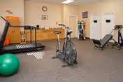 A fitness room in a senior living facility featuring exercise equipment including a treadmill, stationary bike, weight machine, and a green exercise ball. The room has beige walls, a clock, and cabinets along one wall.
