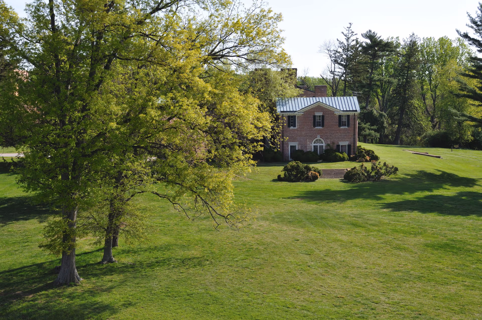 A large green lawn with several trees and a two-story brick house with a metal roof in the background, surrounded by more trees under a clear sky.