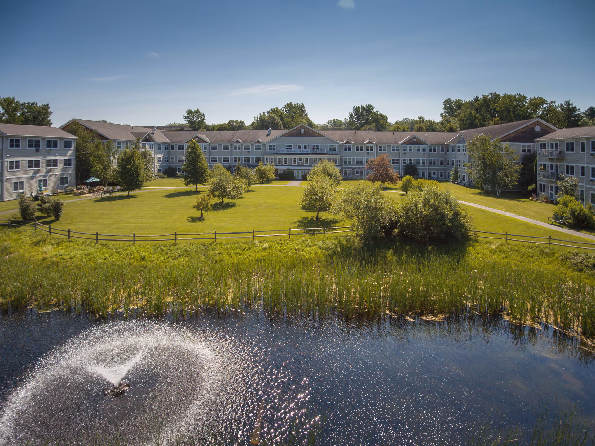 A large senior living facility building with multiple windows and a well-maintained lawn with several trees. In the foreground, there is a pond with a water fountain spraying water upwards. The sky is clear and blue.