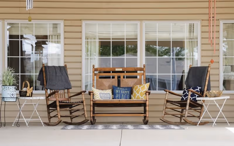 A cozy outdoor seating area with two wooden rocking chairs and a wooden bench swing placed on a porch. The bench swing has decorative pillows with various patterns and texts, including one that says 'This is my happy place.' There are small white side tables next to each rocking chair, one holding a potted plant and the other holding a decorative bowl. The background shows beige siding and large windows with white frames.