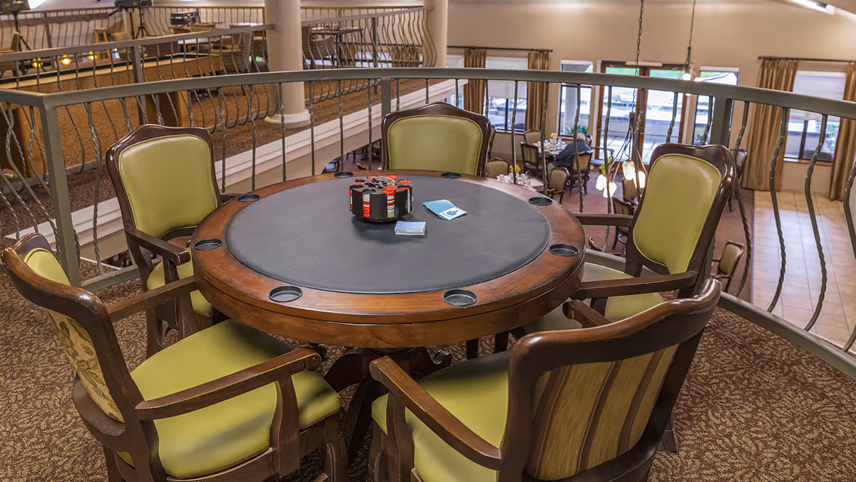 A round poker table with a black playing surface and wooden edges surrounded by five green cushioned chairs. The table has poker chips and cards on it. The setting is an upper-level indoor area overlooking a dining space with large windows and beige curtains.