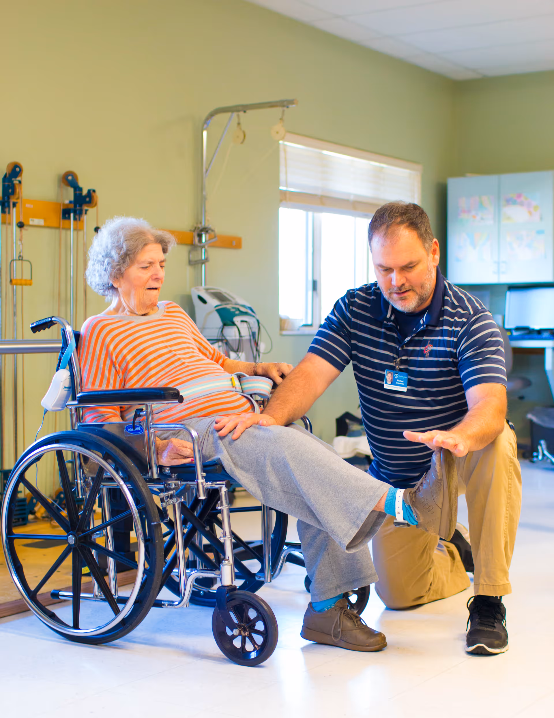 An elderly woman in a wheelchair is assisted by a male caregiver who is kneeling and helping her stretch her leg in a rehabilitation or therapy room with exercise equipment and a window in the background.