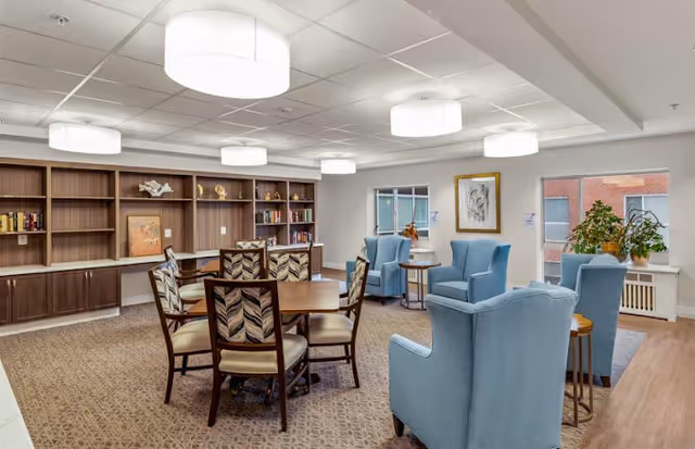 A well-lit common area in Atrium Village featuring a round wooden table with six patterned chairs, blue upholstered armchairs arranged near windows, built-in wooden bookshelves with decorative items, and large ceiling lights.