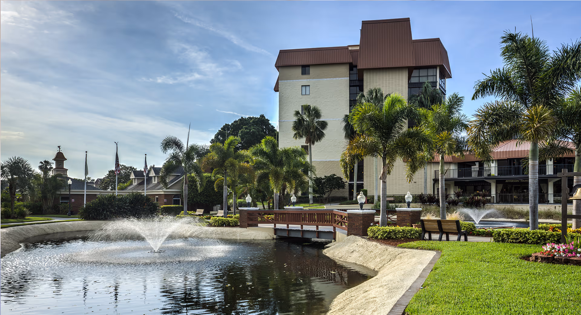 Outdoor view of The Inn at Freedom Square featuring a pond with two water fountains, a small wooden bridge, palm trees, benches, and a multi-story building in the background under a partly cloudy sky.