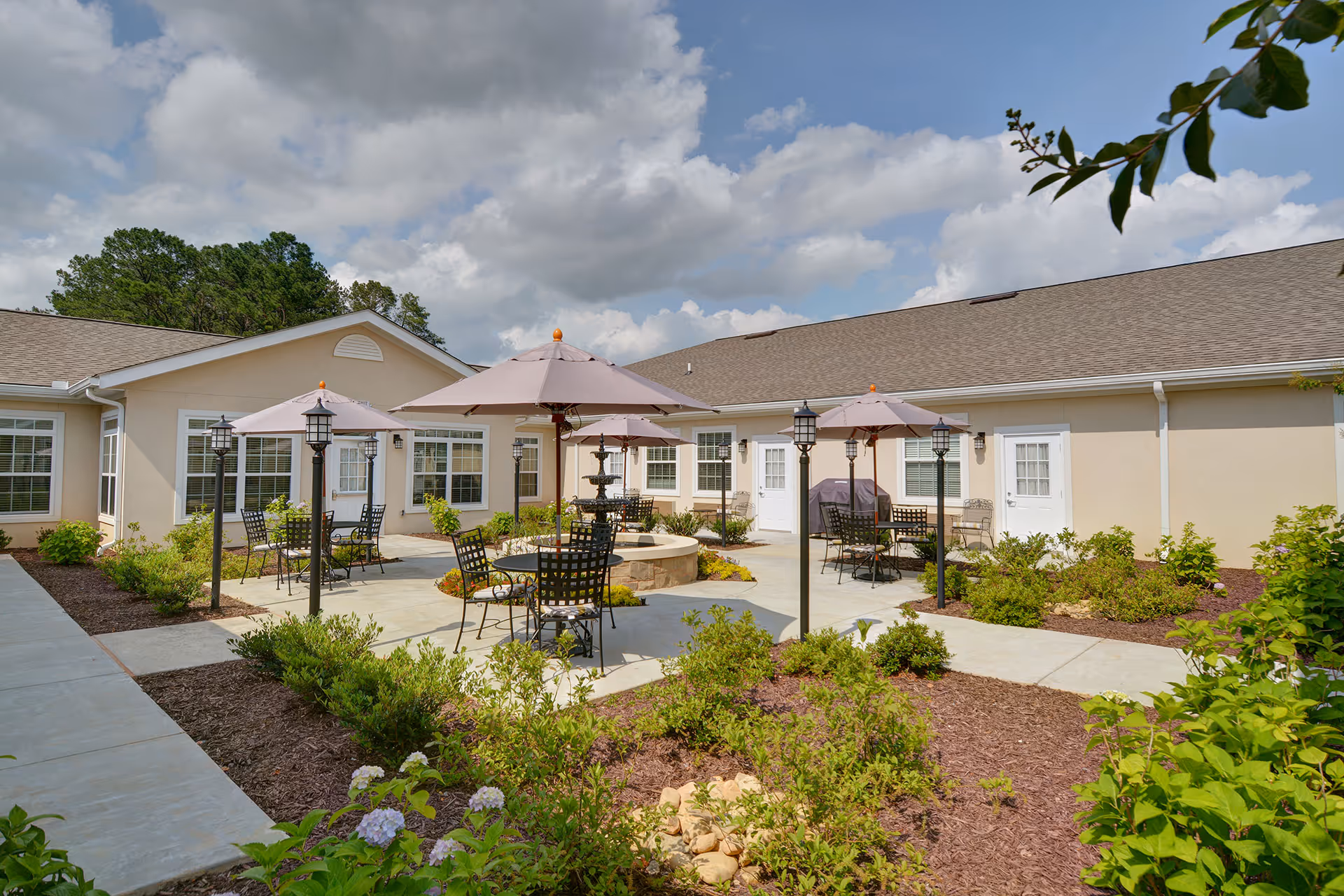 Outdoor patio area at Benton Manor Of Stockbridge with several tables and chairs under umbrellas, a central fire pit, surrounded by landscaped garden beds and shrubs, with a beige building in the background under a partly cloudy sky.