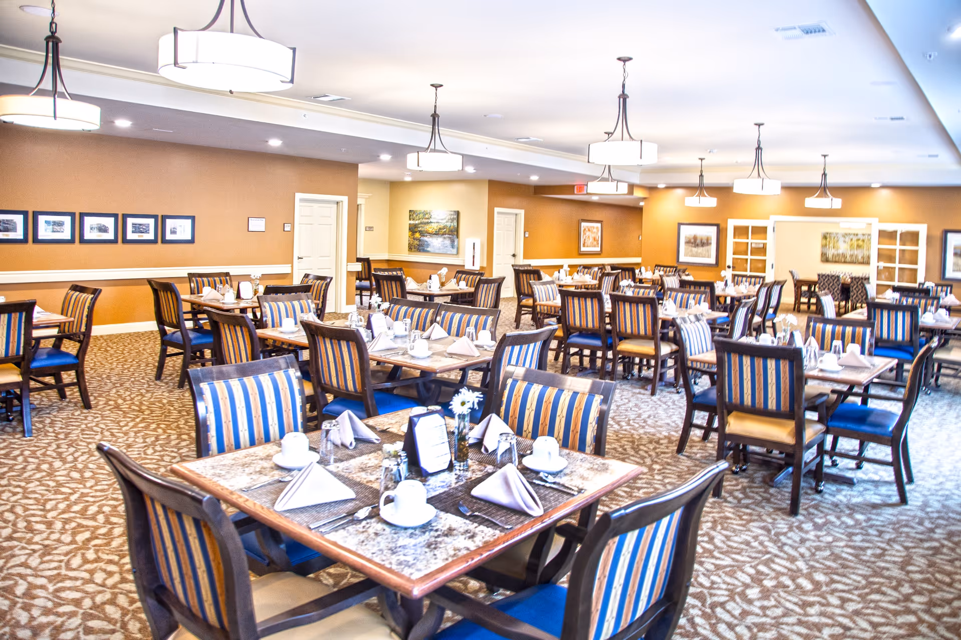 A spacious dining room in a senior living facility with multiple tables set for meals. Each table has place settings including cups, napkins, and utensils, and the chairs have striped upholstery. The room has warm beige walls, carpeted floors with a patterned design, and several ceiling lights providing bright illumination. Artwork and framed pictures decorate the walls.