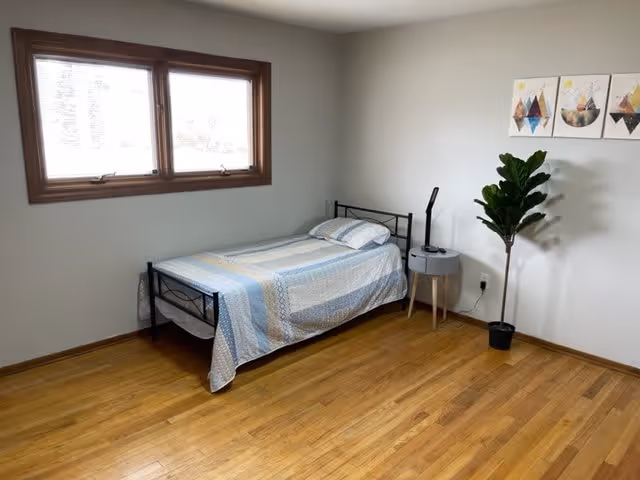 A simple bedroom with a single bed covered with a blue and white patterned bedspread, a small round gray bedside table with a black lamp, a potted plant, three framed abstract art pieces on the wall, and a window with wooden trim letting in natural light.