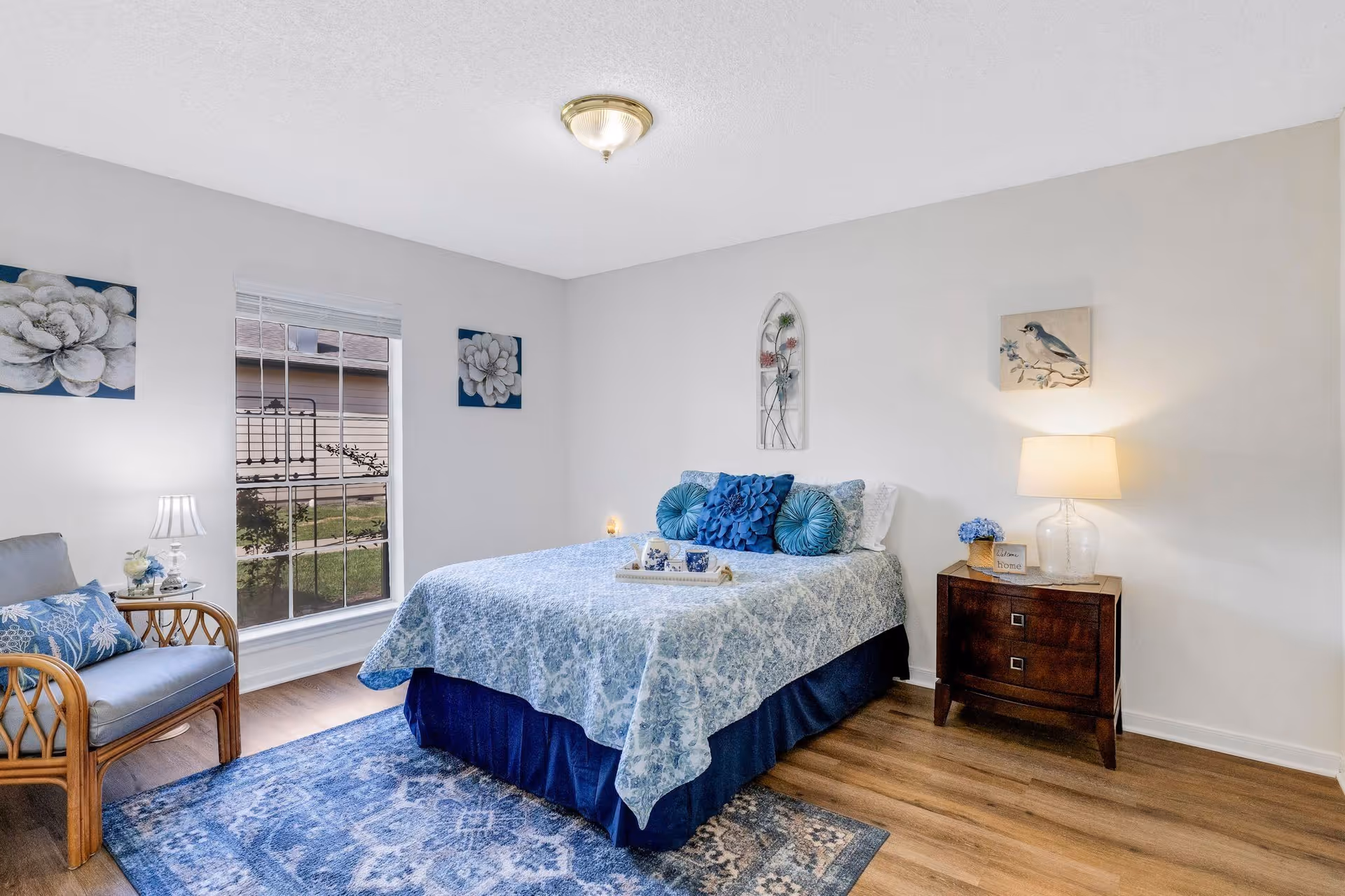 Well-lit bedroom with a made bed in blue floral bedding, a wooden nightstand and lamp, a chair by the window, and decorative wall art.