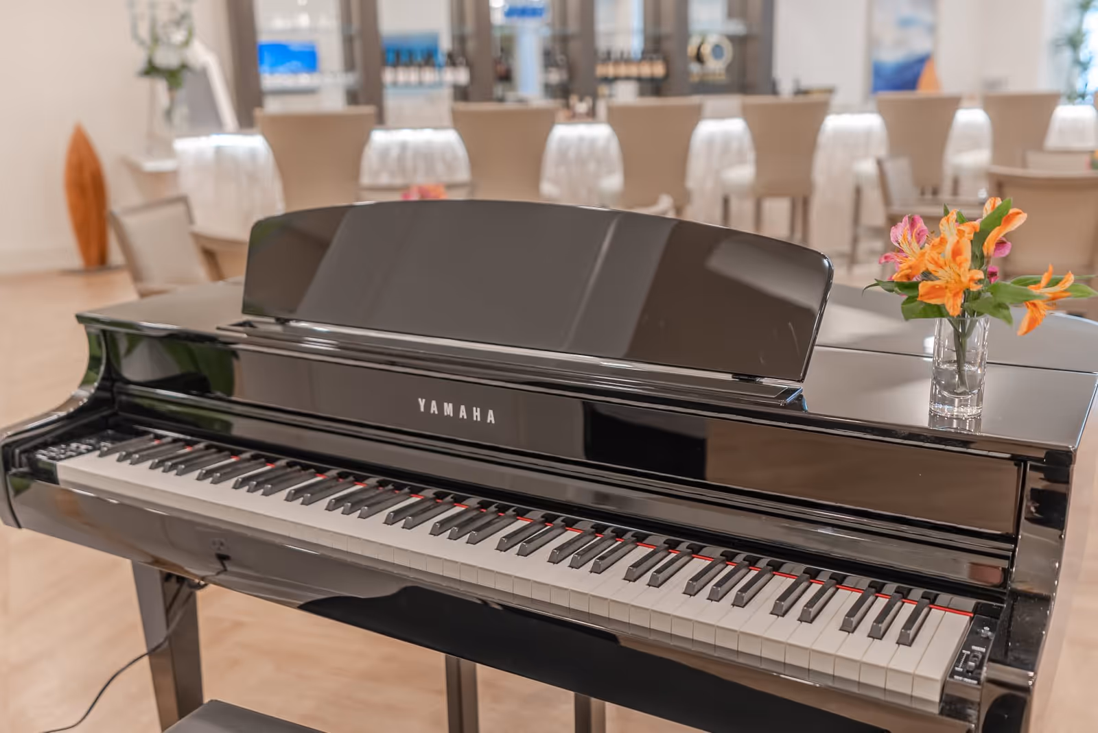 A black Yamaha grand piano with a small vase of orange and pink flowers on top, set in a bright room with beige chairs and tables in the background.