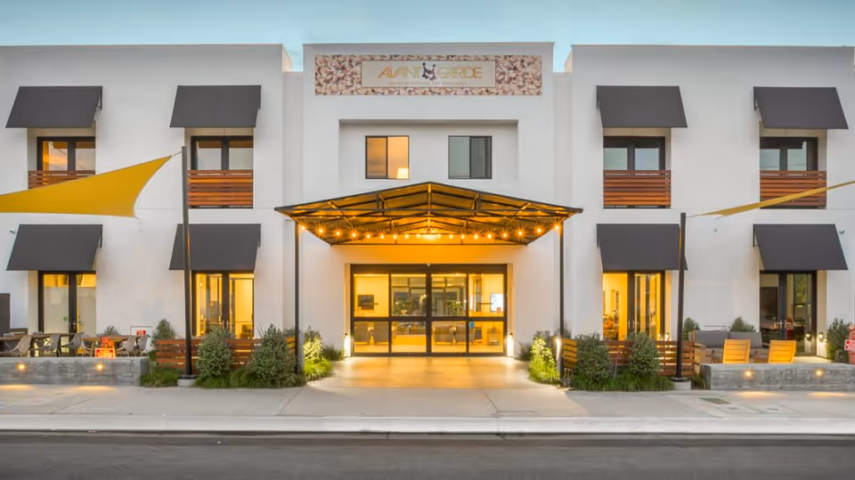 Front entrance of a modern assisted living building with a lit covered entryway, balconies, and outdoor seating at dusk.