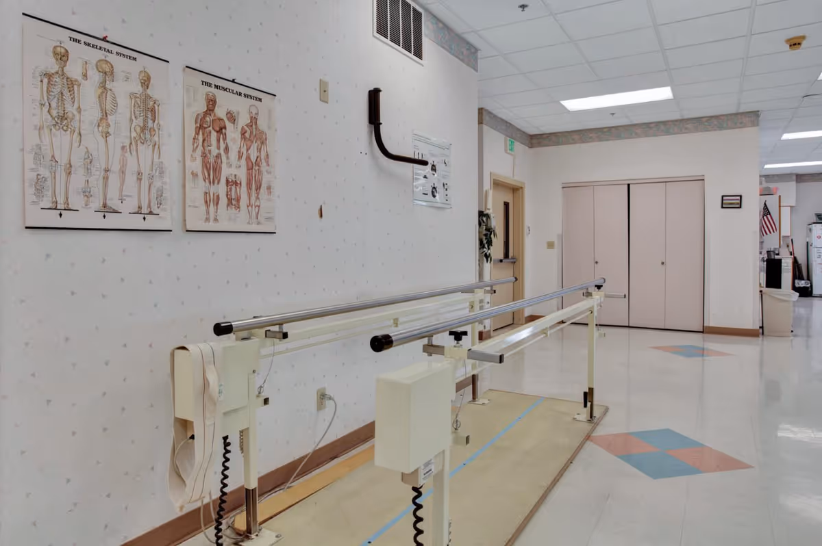 A rehab hallway with parallel walking bars, anatomical charts on the wall, and closed double doors at the far end.