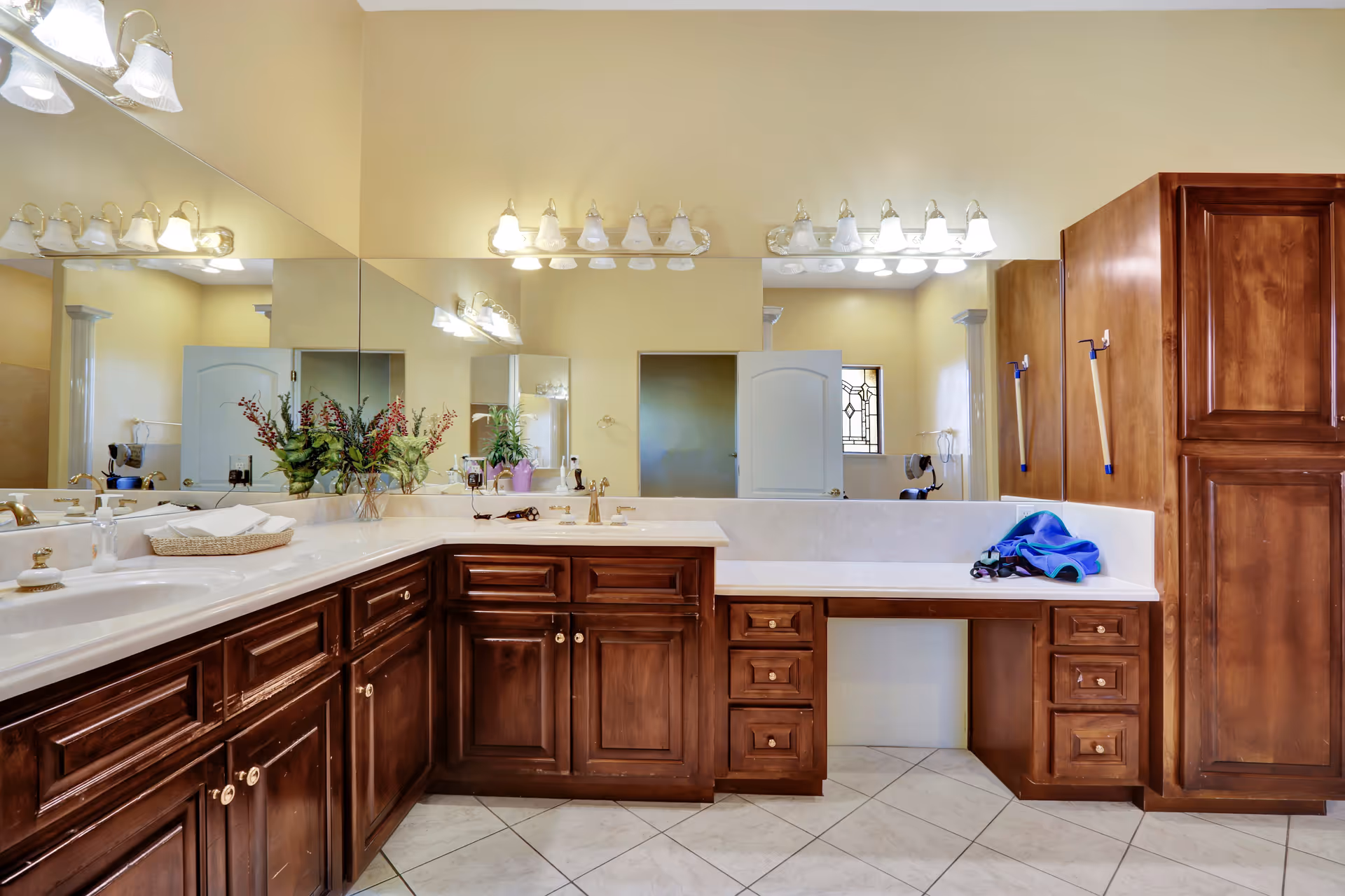 A spacious bathroom with a large L-shaped wooden vanity featuring multiple drawers and cabinets. The countertop is white with two sinks, and there are decorative plants and toiletries placed on it. Above the vanity is a large mirror spanning the length of the counter, with two sets of light fixtures mounted on the wall. The floor is tiled with light-colored tiles, and there is a door and a window with decorative glass in the background.